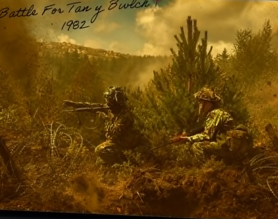 Two camouflaged figures lie in low scrub behind a coil of barbed wire on a hillside of young conifers, one behind a bipod-mounted machine gun, the other with a rifle.